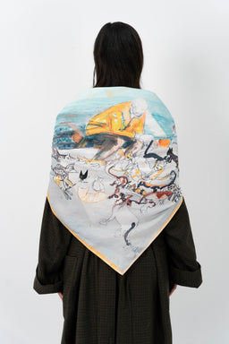 - What the product is: A square scarf featuring an illustration of a person in a yellow top with stray cats, shown draped over the shoulders of a model.
- Apparent audience: Unisex (shown on a woman, but styling and size suggest it can be worn by any gender).
- Notable attributes: Light, pastel-toned color palette with blues and pale yellows; illustrative artwork depicting cats in a seaside or kitchy scene; silk-like sheen with a soft, smooth fabric; edges finished with a narrow border. The scarf is worn draped over the shoulders, visible is the back view of the model.