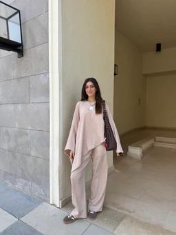 Product is a coordinated two-piece loungewear set in a soft beige color. The outfit appears to be designed for women, featuring a loose, flowing top with wide sleeves and a draped, asymmetrical hem paired with wide-leg pants. The fabric looks crinkled with a gentle wave texture, and the look is completed with a simple necklace and casual shoes. Notable attributes: beige hue, wave/crinkled texture, oversized relaxed fit, matching top and pants set, unstructured silhouette. The model carries a bag and wears neutral-toned footwear, emphasizing a casual, comfortable style.