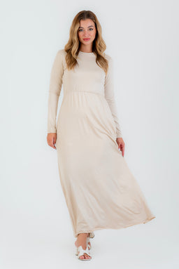 - The photo shows a full-length view of a light beige, long-sleeve, maxi maternity-style dress with a gentle gathered waist. It appears to be made of a soft, natural viscose fabric. The model presents a modest, unisex-leaning fall of the dress with pockets, in a neutral beige tone, paired with light-colored sandals.