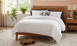 - What the product is: A 3-piece white quilted bedding set laid on a wooden bed, including a comforter/double cover and matching pillow shams.
- Intended audience: Unisex (appears suitable for a bedroom setup with a neutral, coastal-inspired style).
- Notable attributes visible: White quilted fabric with a subtle diamond/textured pattern, navy and white accent pillows, light wood bed frame, cozy living room setting with natural light. Material appears to be soft quilted cotton or a cotton-blend; overall style is clean, simple, and neutral.
