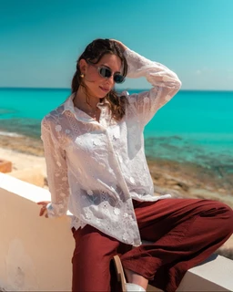 - What it is: A white embroidered blouse with a relaxed fit, sheer lace-like fabric, and long sleeves.
- Intended audience: Women (adult female) based on styling and fit.
- Notable attributes: White eyelet/embroidered fabric, button-front shirt design, classic collar, sheer texture, and lightweight, flowy silhouette paired with burgundy wide-leg pants in a sunny beach setting. No brands shown.