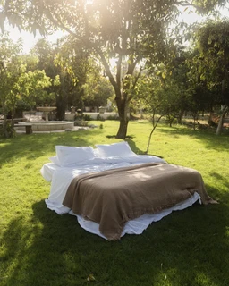 - What the product is: A bed setup featuring a white bed linen set with a brown/mocca throw or blanket layered on top, showcased outdoors on a grassy lawn with trees in the background.
- Intended audience: Unisex (the styling is neutral and could appeal to both men and women; no specific gendered cues).
- Notable attributes visible: White sheets and pillowcases, a brown/beige muslin or soft throw covering the lower half of the bed, outdoor garden setting, daytime lighting, overall casual, airy look. Material emphasis suggests a light, breathable fabric (muslin) with a layered bedding aesthetic.