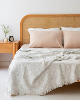 - What the product is: A bed with a light, crinkled muslin bedspread draped over it, featuring a soft, airy texture with a subtle fringe along the edge.
- Apparent audience: Unisex (suits a bedroom setting that both men and women could use).
- Notable attributes: Material appears to be thin, breathable muslin in a pale, neutral color; light gray or off-white tone with a delicate, casual drape; paired with peach-tinted pillows and a wood headboard with woven rattan insert; simple, minimalist bedroom aesthetic.