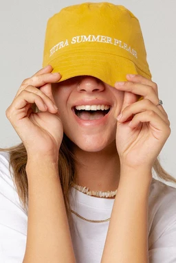 - What the product is: A bright yellow bucket hat with a curved brim.
- Apparent audience: Unisex or women, judging by the model and styling.
- Notable attributes: Lightweight fabric, white embroidered text around the front band reading “EXTRA SUMMER PLEASE,” casual bucket hat style, solid yellow color. Visible pairing with a white top and simple jewelry for a sunny, playful look.