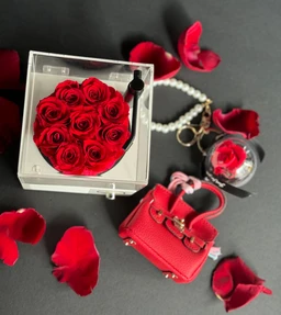 - What the product is: A clear square display box containing a bouquet of red mini roses, paired with a small red handbag and a pearl-adorned keychain, all arranged as a compact gift set. The visible contents also include a tiny round ornament with red rose accents and a chain, suggesting a coordinated accessory set.
- Appereant audience and styling: Appears geared toward adults (unisex accessories with a feminine styling), suggested by the delicate pearl chain, red faux-leather handbag, and decorative mini rose elements.
- Notable material, colour, and style attributes: Red artificial roses sealed in a clear acrylic box; red faux-leather mini handbag with metallic hardware; pearl-studded chain/necklace and a small round pendant with rose motif. Overall color palette is red and white/clear, with a romantic, gift-ready presentation.