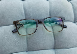 - A pair of rectangular, dark-framed eyeglasses with blue-to-green tinted lenses, shown resting on a light blue quilted surface.
- Intended for adult wearers (unisex appearance based on neutral styling). Notable attributes: anti-blue light coating, plastic/acetate frame with slim temples, subtle gradient lens tint, and a contemporary, square/rectangular frame shape. The photo focuses on the front view with the arms extended.