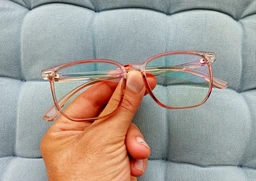 - What the product is: A pair of eyeglasses with a translucent pink/rose-gold frame, held by a hand against a cushioned blue background.
- Apparent audience: Unisex (suitable for men or women), based on neutral styling and frame shape.
- Notable attributes: Rimless or semi-rim design with a thin metal frame, light pink/rose-gold color, clear lenses, straight temples with plastic tips. The photo emphasizes a lightweight, minimal frame suitable for computer use (consistent with “anti blue light” claim).
