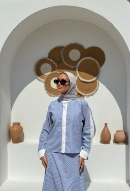 - What the product is: A light blue and white striped long-sleeve shirt with a stand-up placket and white cuffs, shown as a coordinated top paired with matching striped skirt.  
- Apparent intended audience: Women (model wearing a hijab, feminine styling, and tailoring suitable for women's fashion).  
- Notable attributes visible: Blue and white vertical stripe pattern, white placket and cuffs, relaxed fit with a modest design, long sleeves, collarless stand-up style, paired with a matching striped bottom.