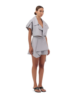 - A light gray, layered skort with a modern, architectural silhouette. The outfit features a short overlayer that forms a wide, draped lapel and a structured, asymmetrical short underneath, creating a hybrid look between a vest-like top and a skirt/shorts combo.
- Audience: women's.
- Notable attributes: light gray color, soft drape with oversized upper panels, cropped top with wide lapels, integrated waistband, asymmetric hem detail, and a matching short underlayer. Material appears structured yet lightweight, with a tonal, monochrome styling. Base footwear: flat black sandals.