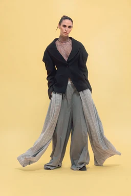 - Product: Mismatched double pants outfit featuring a layered look with wide, flowing drapes attached at the waist and legs.
- Audience: Unisex appearance, but styling reads as fashion-forward adult ensemble (likely marketed to women or gender-fluid audiences).
- Materials/Color/Style: Visible materials include a sheer or lace top under a structured black blazer; outer garments are wide-leg, draped gray pants with additional long, sheer striped panels creating a cape-like overlay. Colors are muted gray and black with a pale, neutral background. Overall style is avant-garde, oversized, and dramatic with a couture-inspired silhouette.
