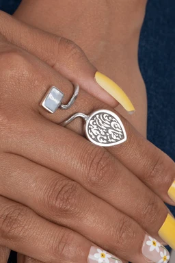 - A silver-tone ring set featuring a square, pale iridescent stone on one end and a round, engraved charm with Arabic-style script on the other end.
- Appears designed for women, shown on a female hand with yellow manicured nails; open, adjustable band with a modern, geometric look.
- Notable attributes include the cool silver metal, light prism stone, and the decorative circular charm with intricate script, all in a contemporary, statement-style ring.