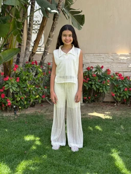 - Description: A girl model wearing an off-white, open-weave two-piece outfit consisting of a sleeveless collared top and wide-leg pants with an airy, sheer texture.
- Audience, styling cues: Appears to target girls or unisex children; light, breezy summer style with a relaxed, resort-inspired vibe.
- Notable materials and attributes: Open-work knit fabric in an off-white color, sheer texture, sleeveless polo-style top, full-length wide-leg pants with elastic or loose fit at the ankles, coordinated set.