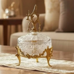 - This is a decorative crystal candy/jewelry dish with a brass base and lid, featuring an ornate finial on top and three gold-toned legs.
- Intended for a general audience (unisex) based on its classic, elegant styling suitable for living room or display use.
- Notable visible attributes: clear cut-glass/crystal bowl with a textured pattern, brass/gold-toned metal base and lid ornament, three curved legs with animal-like motifs, and a matching ornate finial handle on the lid. Color palette is clear crystal with antique gold/brass accents.