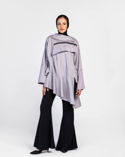 - A loose, light grey poncho-style shirt with an asymmetrical hem and a high collar, worn over black flared pants. 
- Appears intended for women based on the draped silhouette and styling; features a diagonal zip detail across the chest and a layered, cape-like overlay. 
- Notable materials/colors/styles: light grey (poplin-like fabric) with a matte finish, black bottom pants, oversized long sleeves, and an architectural, modernist shirt/shoirt-with-zip accents.