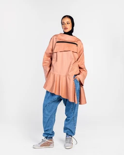 - A peach/soft coral oversized shirt with a high-low hem and asymmetrical peplum-like overlay. It features a high neckline and a detachable or layered front panel with a zippered horizontal chest detail.
- Intended audience appears to be adults (unisex-leaning toward women) based on the styling, relaxed fit, and cropped peplum silhouette.
- Notable attributes: peach color, poplin fabric, long sleeves, front zipper accents, layered flounce/overlay at the chest and hem, paired with loose blue jeans in the image.