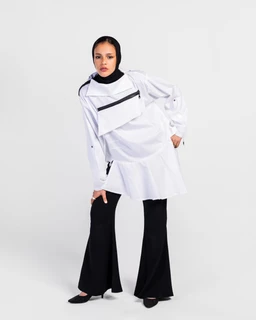 This is a long-sleeve oversized poplin shirt-dress in white with a prominent diagonal zipper across the chest and a high-low hem. The model appears as a unisex or androgynous styling, with a black hijab and black wide-leg pants for contrast, suggesting a modern streetwear vibe. Notable materials and features include crisp white poplin fabric, a visible black zippered chest pocket/zipper detail, and a voluminous, layered silhouette with extended cuffs and a structured, oversized fit.
