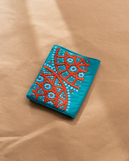 - A teal notebook cover with intricate orange and white embroidered geometric patterns along one corner. 
- Appears to be a unisex accessory, shown in a compact, rectangular notebook style. 
- Visible materials include a fabric cover with colorful embroidery and a soft, woven texture; the overall look is bright, decorative, and handmade-inspired.