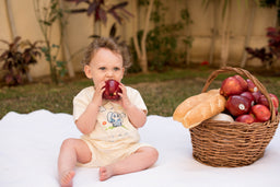 - The image shows a baby sitting on a light-colored blanket outdoors, biting into a red apple. A wicker basket filled with apples and a loaf of bread sits nearby.
- Intended audience: babies/infants (evident from the toddler in the photo; product title suggests “Mommy's Little Peanut” for infants or early walkers, with a unisex presentation in soft, light clothing).
- Visible attributes: light cream onesie, short-sleeve design; natural-toned white blanket; wicker basket; red apples; bread loaf; outdoor garden backdrop. Material emphasis: soft fabric clothing, woven basket, and fresh fruit. Style: candid outdoor baby lifestyle shot with a neutral, gentle color palette.