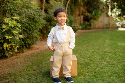 - A young boy model wearing a beige salopette with a white long-sleeve shirt and light tan suspenders, paired with a matching bow tie. He stands on grass in a garden setting, with a wooden block prop behind him and navy shoes.

- Intended audience: boys'. Notable attributes: beige/khaki salopette with suspenders, white shirt, beige bow tie, light tan color palette, casual-formal style, navy shoes.