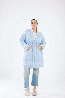 This is a light blue quilted knee-length coat with a soft, plush look. The model appears to be a woman, wearing the coat belted at the waist, with hands in pockets, styling a relaxed, casual vibe. Notable features visible: light baby blue color, padded quilted panels, belt-tied waist, and a cozy, oversized fit with cropped sleeves, paired with light denim jeans and sneakers. Material suggests a fluffy, insulating fabric (quilted/synthetic filling) and a smooth outer shell.
