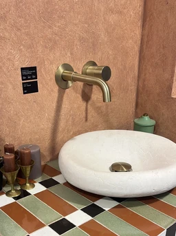 - What it is: A wall-mounted faucet and a round stone sink on a tiled counter, shown in a bathroom setting.
- Intended audience: Unisex (based on neutral design and faucet/sink styling suitable for both genders).
- Notable attributes: The sink is a smooth, white stone or stone-like basin with a brushed metal faucet in a warm brass/bronze tone. The surrounding decor includes multicolored ceramic tiles (green, brown, black, white) and matching accessories, conveying a rustic-modern style.