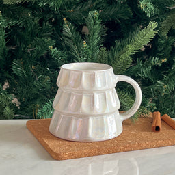A white iridescent Christmas mug with a tiered, ruffled stack design and a single handle. It appears to be a handmade ceramic mug, shown on a cork board with evergreen branches in the background, suggesting a festive holiday aesthetic. Intended audience seems unisex; the visible style emphasizes a chunky, stacked look with a glossy, pearlescent glaze in white. Notable attributes: handmade ceramic, iridescent white glaze, tiered/ringed geometric pattern, wide mouth, side handle.