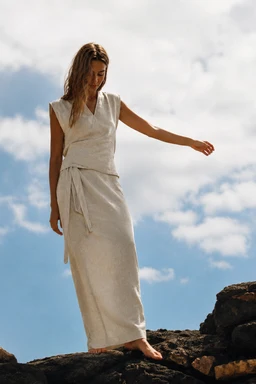 - What the product is: A sleeveless linen wrap dress or robe-style set, shown as a two-piece lounge/robe with a tied waist against a rocky outdoor backdrop.
- Intended audience: Women (based on the model and styling).
- Notable attributes visible: Made of light, natural linen fabric in a soft off-white or cream color; sleeveless V-neck top with a wrap/overlayer and a long, ankle-length skirt with a tied belt at the waist; relaxed, flowy silhouette with a simple, minimalist style suitable for loungewear or resort wear.