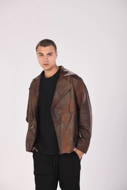 A vintage-style leather jacket with a slightly oversized, relaxed fit. The jacket is brown distressed leather, with wide lapels and a draped, open-front design. The model appears to be a unisex presentation (androgynous fit), paired with a black shirt and black pants to emphasize the leather texture. Visible attributes include the brown patina leather, asymmetric front with wide notched lapels, and a casual, slightly cropped length.