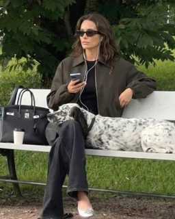 - Product: A casual, on-the-go outfit styled for a relaxed day off. The look is modeled by a woman sitting on a bench with a dog beside her, using a phone and wearing portable earphones.
- Audience: Women's/mixed unisex casual wear inferred from the model and styling.
- Materials and style: Olive/brown bomber-style jacket over a dark top, loose dark jeans, white sneakers, and a simple necklace. Accessories include a black handbag and sunglasses, with a coffee cup on the bench. Overall vibe is laid-back, urban-casual.