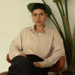 A beige linen shirt worn by a seated female model. The shirt has a classic collar and button-front placket, with long sleeves and a relaxed fit. The fabric appears lightweight and slightly textured, in a soft, natural beige hue, paired with dark trousers and simple accessories for a minimalist, unisex to feminine look.