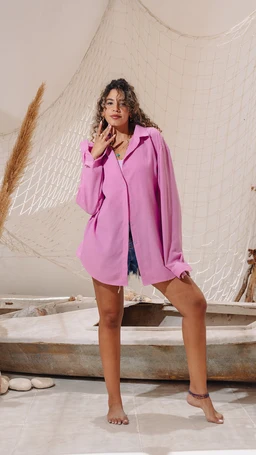Description: A woman models an oversized pink shirt, worn as a lightweight outer layer or shirt-dress. Audience: unisex or women’s (based on styling and fit). Notable attributes: bright pink linen-like fabric, relaxed loose-fit silhouette, long sleeves with cuffs, drop-shoulder design, collar and button-front opening, shown with denim shorts underneath in a casual, beachy setting.