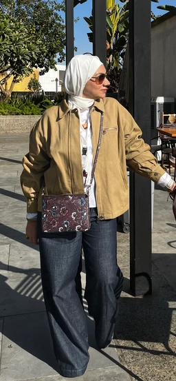 - What the product is: A light tan/brown zip-front jacket worn by a model outdoors.
- Apparent audience: Women (styled with a hijab and feminine accessories; full-length dark jeans and crossbody bag suggest a women's fashion item).
- Notable visible attributes: Light jacket in a camel/tawn shade with zip front and fitted cuffs; white inner shirt visible at neckline and sleeves; dark wide-leg jeans; patterned crossbody bag; white hijab and sunglasses. Material appears to be a lightweight, possibly cotton or lightweight denim-like fabric; overall styling is casual, street-ready.