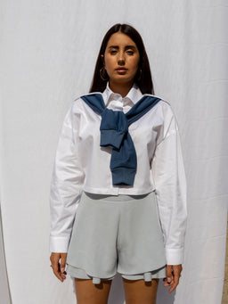 - The image shows a woman wearing a white long-sleeve button-down shirt with a blue sweater tied around the shoulders. A navy blue top is draped and knotted at the front, creating a layered, preppy look.
- Intended audience: women's fashion.
- Notable attributes: crisp white cotton shirt, light gray high-waisted shorts with a loose, tailored fit, and a navy blue sweater tied around the neck. The overall style is smart-casual with a collegiate, club-style vibe. Material hints include cotton for the shirt and knit for the sweater.