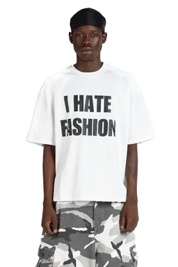 - What it is: A casual men's t-shirt featuring a bold graphic text that reads "I HATE FASHION" in black letters. 
- Apparent audience: Men's; relaxed, oversized fit with dropped shoulders and wide sleeves.
- Notable attributes: White cotton jersey fabric, short sleeves, crew neck, and a loose, boxy silhouette. The model also wears a black durag and gray camo-pattern shorts, but only the T-shirt design is described as the product.