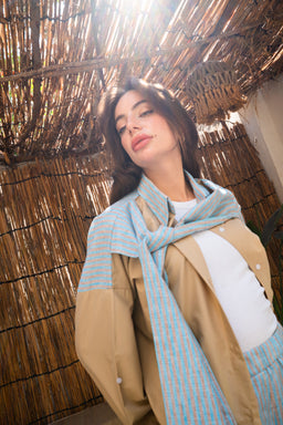 - What the product is: An on-shoulder shirt look featuring a light blue scarf draped over a beige/brown outer garment with a white top underneath.
- Apparent audience and styling: Womenswear, styled with a relaxed, chic, draped look suitable for casual or semi-formal outfits.
- Notable attributes observed: Light blue scarf with a subtle texture wrapped over a tan/taupe coat and white inner top, creating a soft, layered, contemporary aesthetic. The model has a confident pose in an outdoor, sunlit setting with natural woven backdrop.
