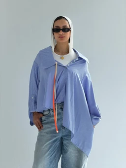 - A light blue oversized striped shirt with a loose, draped silhouette. The model wears it layered over a white hoodie and light-wash jeans, accessorized with a thin belt, black sunglasses, and a white headscarf.
- Audience: primarily designed for women, suggested by the relaxed, oversized fit and styling.
- Notable attributes: striped pattern, oversized draped cut, pale blue color, long sleeves with rolled cuffs, collared neckline, and a casual streetwear vibe. Material appears lightweight cotton or a similar breathable fabric. No branding visible.