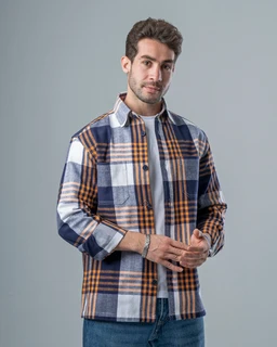 A man models a long-sleeve overshirt with a bold checkered pattern in orange, navy, blue, and white tones. It appears intended for a unisex or men’s audience, worn open or buttoned over a light layer, with a casual, workwear-inspired style. Notable features include the oversized plaid blocks, a collared neckline, button-front closure, and a soft, brushed cotton-like fabric.