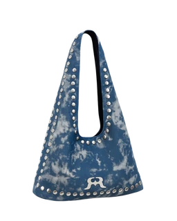 - What the product is: A blue tie-dye denim tote bag with a rounded, slouchy silhouette and long parallel handles.
- Intended audience: Unisex, based on simple, roomy tote styling and neutral denim finish.
- Notable attributes: Indigo-blue tie-dye denim fabric with a row of metal studs along the outer edges and handle opening; a small white emblem/logo near the base edge. No visible branding beyond the emblem.