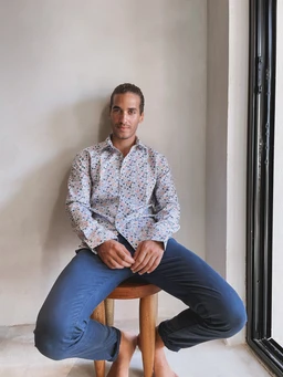 - A male model seated on a wooden stool, wearing a long-sleeve button-up shirt with a small allover print and slim-fit blue pants. The setting is indoors by a large window with natural light. Notable attributes: light-colored shirt with a subtle pattern, navy/blue trousers, and a casual yet put-together look. Audience: men's apparel. Material and style cues: lightweight woven shirt, cotton or cotton-blend, casual dress-casual hybrid style.