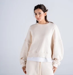 A women's casual sweatshirt featuring a relaxed, oversized silhouette with voluminous sleeves and a ribbed crew neck. The garment layers over a white underlayer hem, creating a two-tone look with a pale beige exterior and a visible white hem peeking at the bottom. Notable materials appear to be a soft knit (likely cotton or cotton-blend) with a plush, comfortable feel, in a neutral beige tone and minimalistic styling.