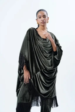 - Product: A draped, loose-fitting poncho-like top or tunic from the Layaly Set.
- Intended audience: Women (adult female model, draped styling and accessories suggest feminine wear).
- Visible attributes: Dark olive to black velvet-like fabric with a soft, flowing drape and fringe trim along the hem and sleeves. The garment features a wide, cowl neckline and voluminous, cape-like silhouette with layered gathers. No branding shown.