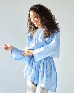 - What the product is: A light blue, loose-fit blouse with a peplum-like waist and wide, long sleeves.
- Intended audience: Women (styled on a female model).
- Notable attributes: Soft, airy blue fabric with a crinkled texture; off-the-shoulder/boat neckline with gathered shoulders; voluminous sleeves and a cinched, flared lower silhouette giving a relaxed, blousey look. No visible branding.