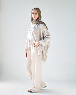 - Description: A beige, modest two-piece outfit featuring a loose, wide-leg pants set with a matching long-sleeve top or poncho-style outer layer. The look is soft and flowy with a relaxed fit.
- Audience and styling cues: Appears designed for women; modeled with a hijab, suggesting a modest, contemporary Middle Eastern/softly traditional aesthetic.
- Materials, color, style notes: Light-beige fabric with a matte finish; outer layer has a draped, cape-like silhouette with subtle floral or star-like embroidery; overall color palette is neutral beige with a slightly darker scarf.