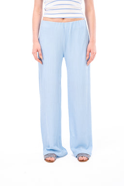 - What it is: Light blue, wide-leg pants with a relaxed fit and an elastic waistband.
- Intended audience: Women, based on styling with a cropped striped top and sandals.
- Notable attributes: Soft, drapey fabric in a pale blue color; full-length, loose silhouette with a gathered/elasticized waistband; casual, lounge/relaxed-fit style.
