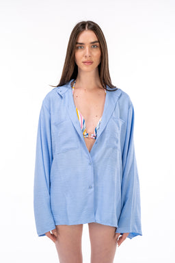 This is a light blue linen shirt with a relaxed, oversized fit and long sleeves, shown unbuttoned over a colorful striped bikini top. The model appears female, suggesting the intended audience is women. Notable details include the breathable linen fabric, pastel blue color, drop shoulders, and a casual, beachy styling.