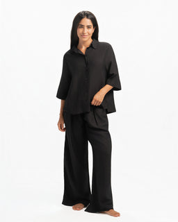 A black linen shirt styled as a relaxed, flowy top with a standard collar and button placket. The model wears it with matching wide-leg pants, giving a coordinated loungewear or casual-smart look; sleeves are three-quarter to mid-length with a loose, draped silhouette. Visible attributes include a soft, breathable linen material, solid black color, and a loose, oversized fit with a high-low hem. Audience appears to be unisex or women based on styling, cut, and model.