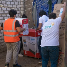 - What the product is: A group of people unloading or handling boxed appliances, including a boxed cooking appliance (likely a stove/oven) and other large home electronics.
- Apparent audience: Unisex (visible people of different ages/genders; focus on general consumer use).
- Notable attributes: Large cardboard and boxed packaging in red/white with appliance imagery; people wearing casual clothing, one person in an orange safety vest. Text and branding unobtrusive; materials are cardboard boxes and printed product images. The scene suggests a home appliance or cookware product distribution, with a prominent stove/oven box visible.