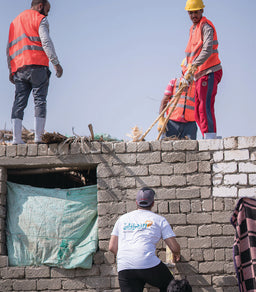 - What the product is: A construction scene showing workers on a brick rooftop wearing safety gear; not a single product, but an overall workwear and safety equipment context.
- Apparent audience: Unisex (men and/or workers) based on the model types and attire.
- Notable attributes visible: High-visibility safety vests (orange), long-sleeve shirts, work pants, hard hats (yellow), work gloves, and rugged outdoor footwear. Built environment includes a brick wall, exposed rebar/ropes, and a blue tarpaulin window cover. The style is practical industrial/workwear suitable for roofing or construction settings.