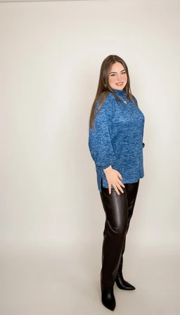 - A blue marbled knit sweater with a high neckline and long sleeves, modeled by a woman. The cut is relaxed with a slightly longer back and side slits. Material appears soft and thick, suggesting a wool blend; color is a heathered blue with darker marbling. The styling is casual yet polished, paired with black leather-look pants and black ankle boots for a women’s audience.