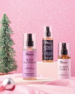 Product appears to be a small gift set of Bath & Body Works-style body mists (The Bathland “Mini Mist” collection). Intended for a general audience (unisex or gifting, with a light, playful unisex to feminine vibe judged from pastel pink background and floral bottle labels). Notable attributes: pastel pink and lavender bottles, spray misters, clear labels with fragrance names like Wild Blossom and Warm Vanilla, set against a pink, festive display with a small Christmas tree.
